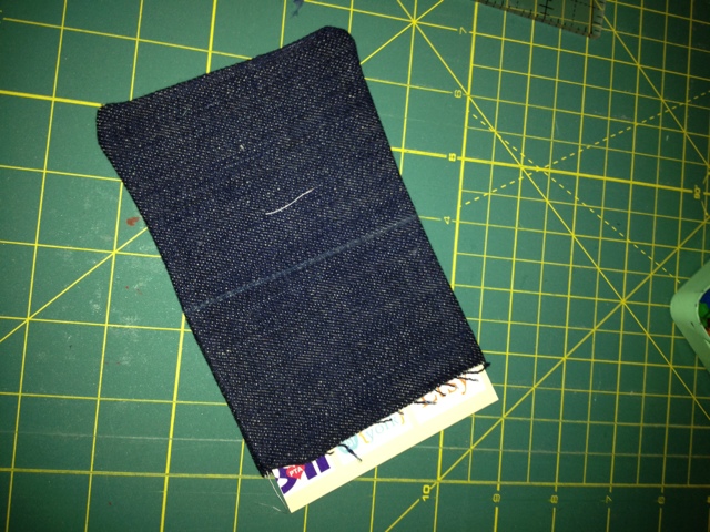 Dark denim fabric pouch lying on a green cutting mat with grid lines, a colorful fabric edge peeking out, neutral crafting mood