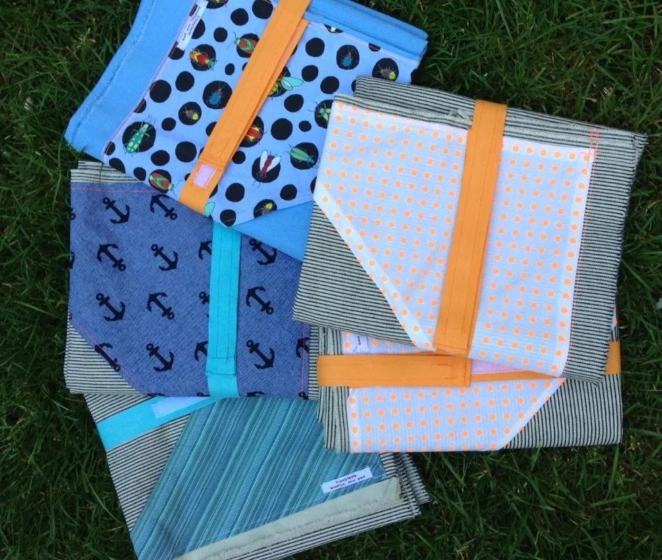 Stack of folded fabric items with polka dot, anchor, and striped patterns in orange and teal, laid casually on green grass