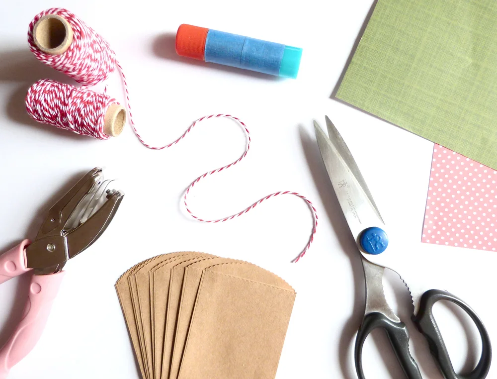 Flat lay of craft supplies including twine spools, a glue stick, scissors, small kraft paper bags, and colored paper squares on a neutral background