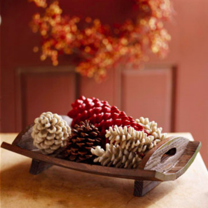 Warm-toned flat lay of decorative pinecones in white, red and brown shades on a wooden tray with a softly blurred autumn wreath in the background, evoking cozy seasonal atmosphere