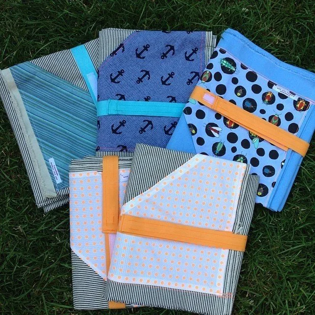 Assorted folded fabric items with striped, polka dot and anchor patterns laid on green grass, colorful casual outdoor mood