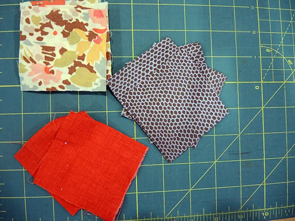 Small stacked fabric squares in floral, dotted, and solid red patterns on a green cutting mat with grid lines, crafting mood