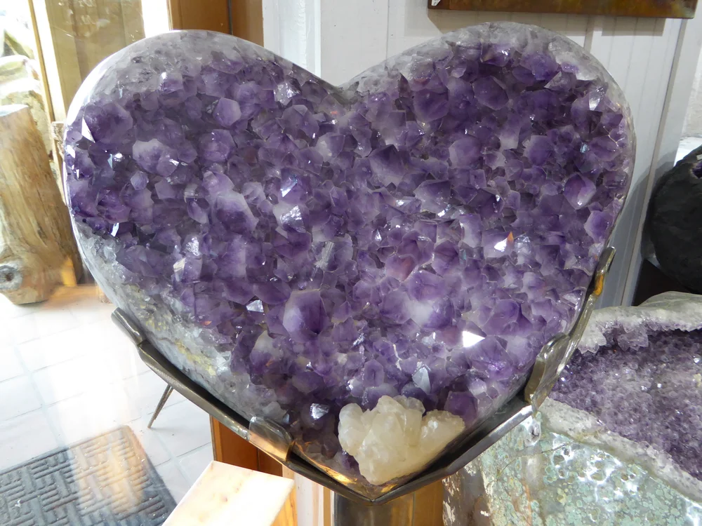 Heart-shaped amethyst crystal cluster with deep purple crystals and a small white calcite accent on a display stand, moody violet and indigo tones